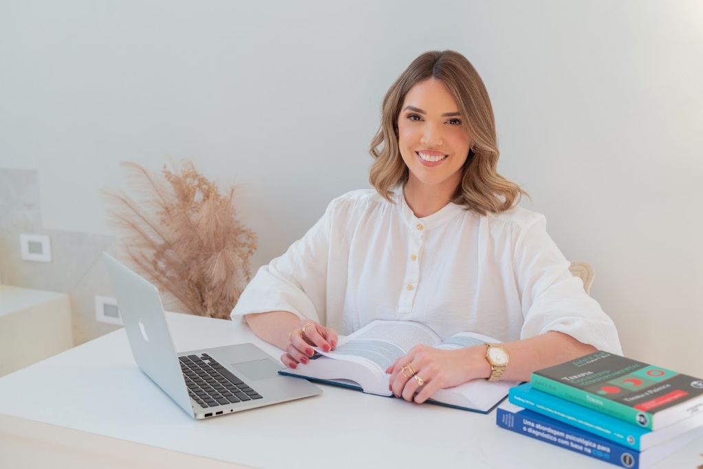 Psicóloga Ana Tolomeu sentada em consultório com livros e notebook